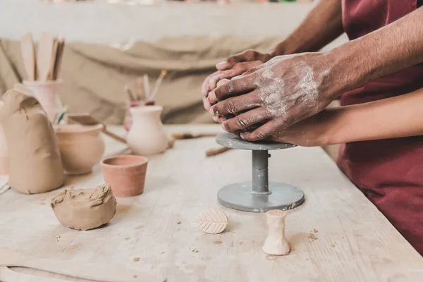 Vue partielle du jeune couple afro-américain sculptant le pot d'argile avec les mains jointes dans la poterie — Photo de stock