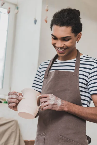 Sonriente joven afroamericano hombre esculpiendo olla de barro con palo en cerámica - foto de stock