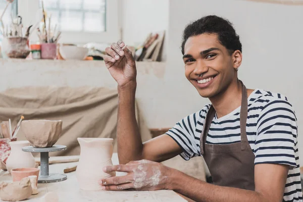 Smiling young african american man looking at camera near clay pot and stick in hand in pottery — Stock Photo