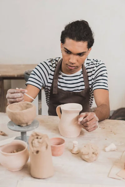 Hombre afroamericano joven serio esculpir olla de arcilla y palo en la mesa con el equipo en cerámica - foto de stock
