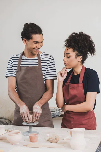 Sorridente jovem afro-americano homem fazendo panela de barro perto de mulher com a mão perto do rosto em cerâmica — Fotografia de Stock