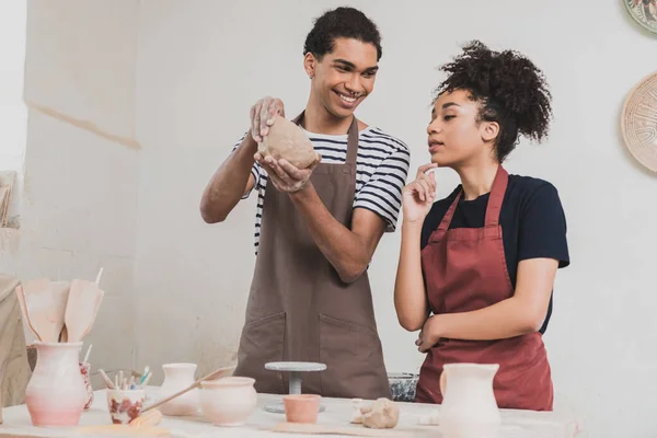 Souriant jeune homme afro-américain faire pot d'argile près de la femme avec la main près du visage en poterie — Photo de stock