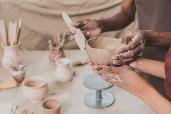 Vista parcial de pareja afroamericana joven haciendo olla de arcilla con espátula y manos en la mesa con equipo en cerámica - foto de stock