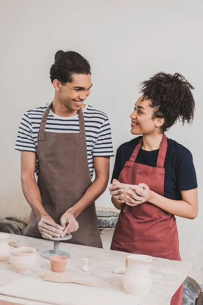 Lächelndes junges afrikanisch-amerikanisches Paar in Schürzen, die Tontöpfe herstellen und einander in Töpfern anschauen — Stockfoto