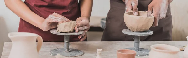 Vue partielle du jeune couple afro-américain dans des tabliers faisant des pots d'argile sur la table avec équipement en poterie, bannière — Photo de stock