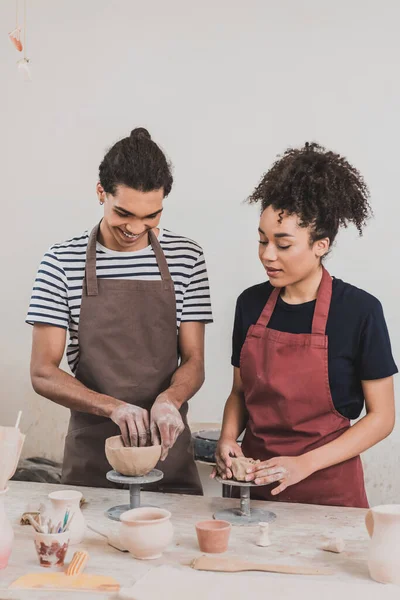 Alegre joven afroamericano pareja en delantales haciendo ollas de barro en la mesa con equipo en cerámica - foto de stock