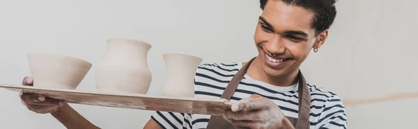 Smiling young african american man looking at clay pots on tray in pottery, banner — Stock Photo