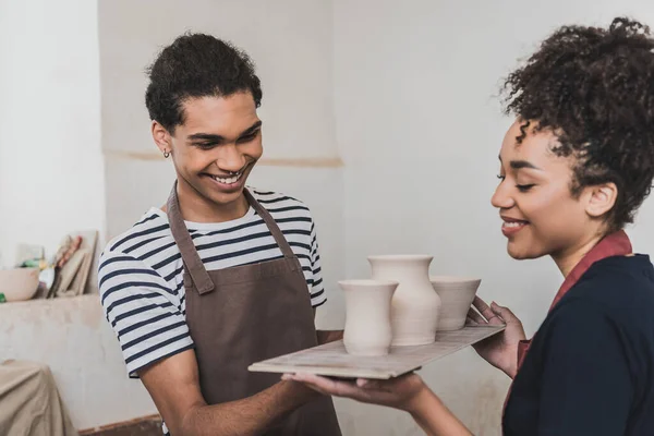 Sonriente pareja afroamericana joven sosteniendo ollas de barro en bandeja en cerámica - foto de stock