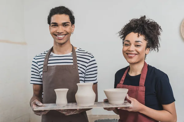 Sonriente joven afroamericano pareja sosteniendo ollas de arcilla en bandeja y mirando a la cámara en cerámica - foto de stock