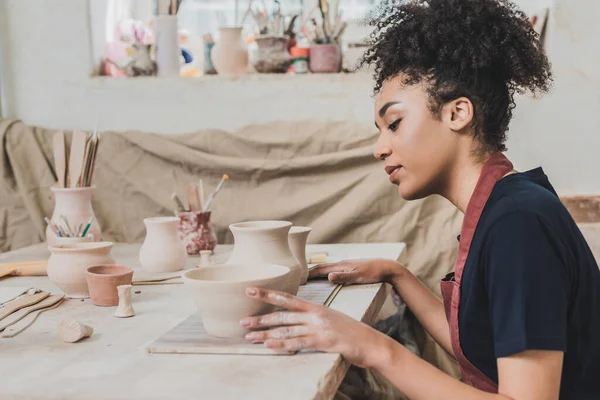 Joven afroamericana enfocada sentada a la mesa con ollas de barro en cerámica - foto de stock