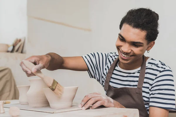 Smiling young african american man glazing clay pots with brush in pottery — Stock Photo