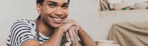 Улыбающийся молодой африканский американец, сидящий с руками возле лица и смотрящий на камеру в керамике, баннере — стоковое фото
