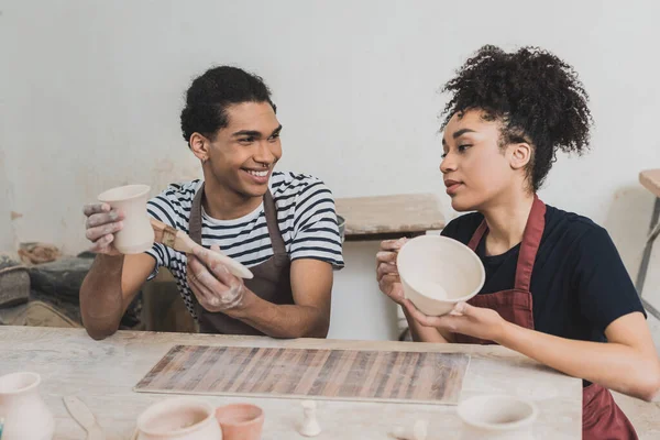 Joven africano americano pareja acristalamiento arcilla ollas con cepillos cerca de mesa con equipo en cerámica - foto de stock
