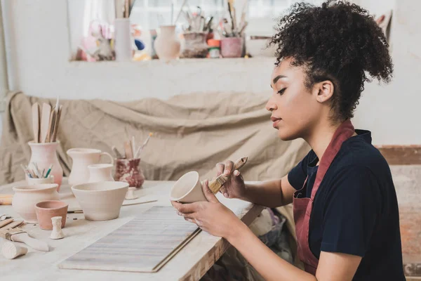 Sérieux jeune femme afro-américaine glaçure pot d'argile avec brosse près de la table avec équipement en poterie — Photo de stock