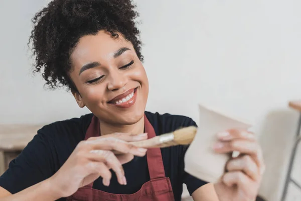 Sonriente joven afroamericana mujer acristalamiento arcilla olla con cepillo en cerámica - foto de stock