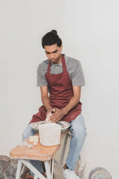 Serio joven afroamericano hombre en ropa casual y delantal modelado de arcilla mojada en la rueda en cerámica - foto de stock