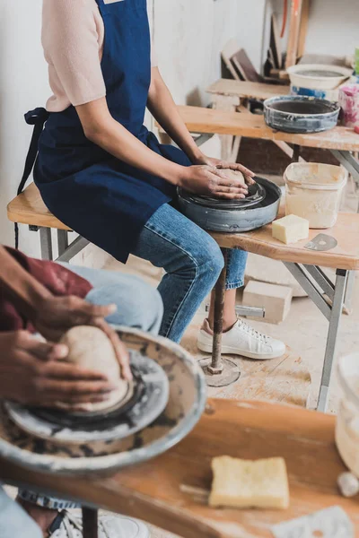 Vue partielle du jeune couple afro-américain en vêtements décontractés et tabliers modelant l'argile humide sur roues en poterie — Photo de stock