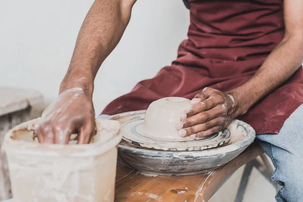 Partial view of young african american man modeling wet clay on wheel and washing hand in plastic box in pottery — Stock Photo