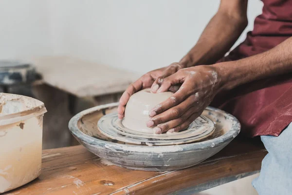 Partial view of young african american man modeling wet clay on wheel with hands in pottery — Stock Photo