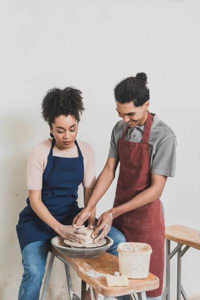 Happy young african american couple modeling wet clay on wheel with hands in pottery — Stock Photo