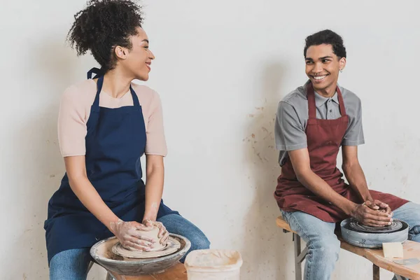 Partial view of young african american couple looking at each other and modeling wet clay on wheels with hands in pottery — Stock Photo