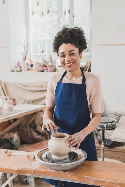 Sonriente joven afroamericana mujer modelando mojado arcilla olla en rueda en cerámica - foto de stock