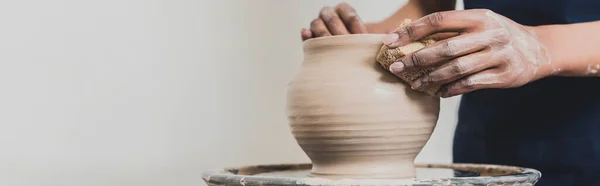 Vue partielle de jeune femme afro-américaine modelant pot d'argile humide sur roue avec éponge en poterie, bannière — Photo de stock