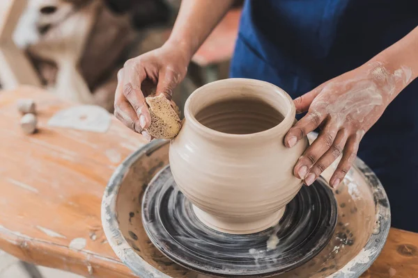 Vue partielle de jeune femme afro-américaine modelant pot d'argile humide sur roue avec éponge en poterie — Photo de stock