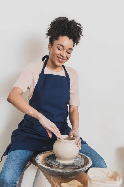 Lächelnde junge afrikanisch-amerikanische Frau in lässiger Kleidung und Schürze, die nassen Tontopf auf einem Rad modelliert, mit Töpferhänden — Stockfoto