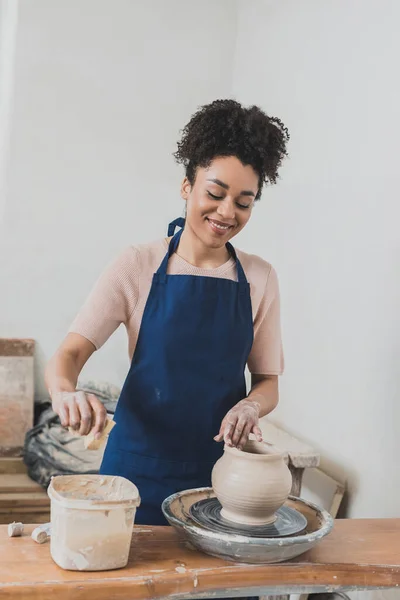 Sonriente joven afroamericana mujer en delantal modelado mojado olla de arcilla en la rueda y apretando la esponja con la mano en cerámica - foto de stock