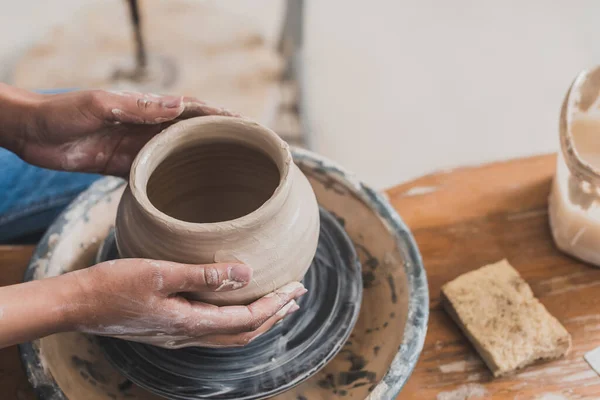 Vista parziale di giovane donna afro americana modellante pentola di argilla bagnata su ruota con mani su panca con spugna in ceramica — Foto stock