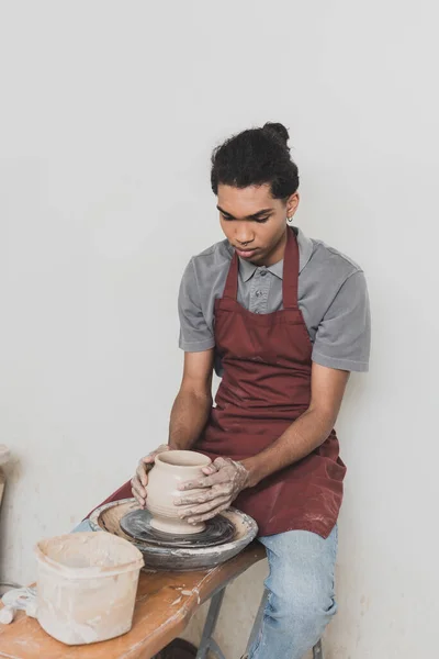 Серьезный молодой африканский американец, моделирующий мокрый глиняный горшок на колесах с руками в керамике — стоковое фото