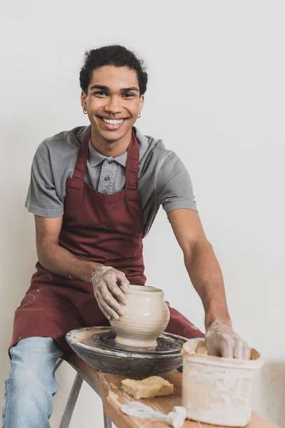 Smiling young african american man modeling wet clay pot on wheel with hands in pottery — Stock Photo