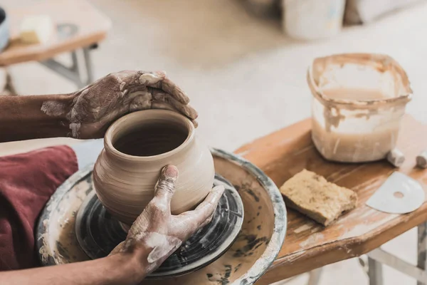 Partial view of young african american man modeling wet clay pot on wheel with hands in pottery — Stock Photo