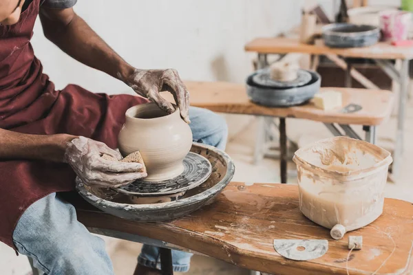 Partial view of young african american man in casual clothes and apron modeling wet clay pot on wheel in pottery — Stock Photo