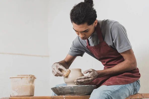 Serious young african american man in casual clothes and apron modeling wet clay pot on wheel in pottery — Stock Photo