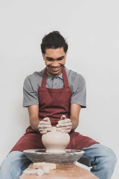 Souriant jeune homme afro-américain en vêtements décontractés et tablier modelage pot d'argile humide sur roue en poterie — Photo de stock