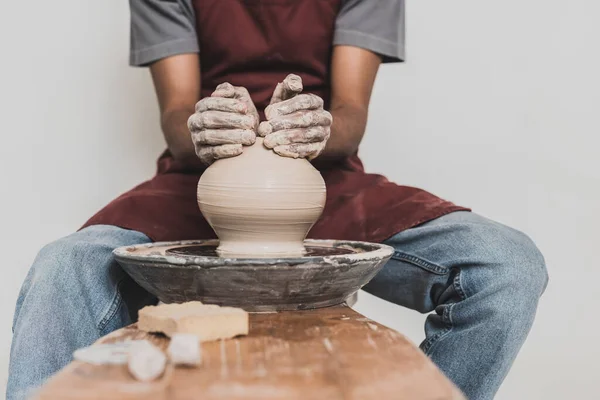 Partial view of young african american man sitting on bench and shaping wet clay pot on wheel with hands in pottery — Stock Photo