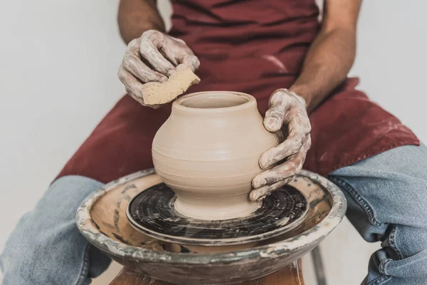 Partial view of young african american man sitting on bench and shaping wet clay pot on wheel with sponge in pottery — Stock Photo