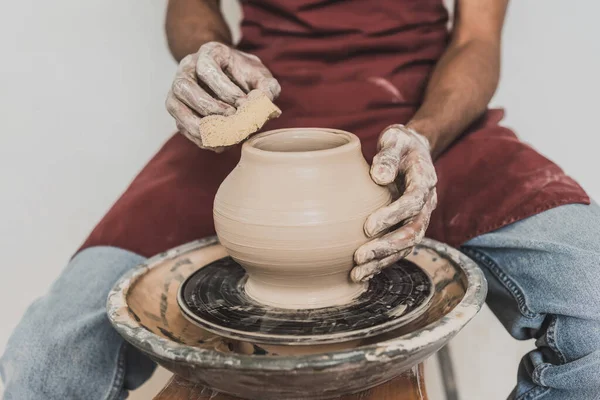 Vista parziale del giovane uomo afroamericano seduto su panchina e modellante pentola di argilla bagnata su ruota con spugna in ceramica — Foto stock