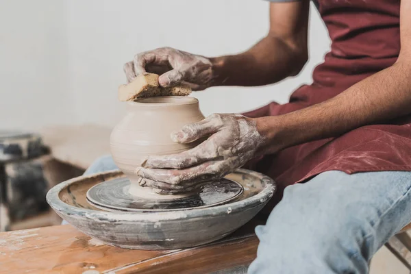 Partial view of young african american man in jeans sitting on bench and shaping wet clay pot on wheel with sponge in pottery — Stock Photo