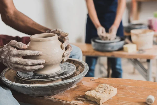 Partial view of young african american man shaping wet clay pot on wheel near blurred woman in pottery — Stock Photo