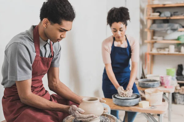 Concentré jeune couple afro-américain en vêtements décontractés et tabliers façonnant des pots d'argile humide en poterie — Photo de stock