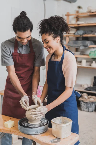 Pareja afroamericana joven positiva en delantales que dan forma a maceta de arcilla húmeda en la rueda con las manos en cerámica - foto de stock