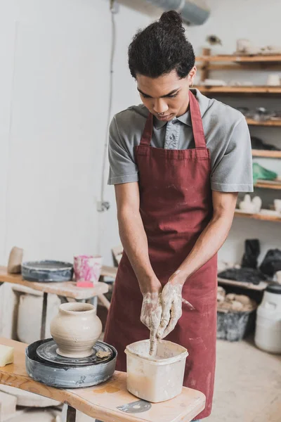 Serious young african american man washing hands in plastic box near wet clay pot on wheel in pottery — Stock Photo