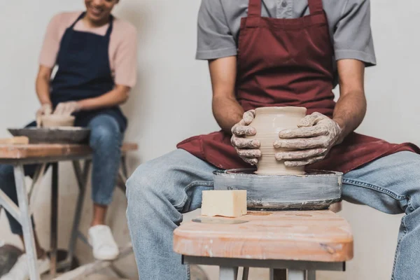 Vista parziale del giovane uomo afro-americano che fa pentola di argilla bagnata sulla ruota vicino alla donna sfocata in ceramica — Foto stock