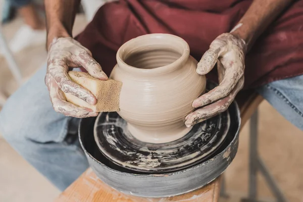 Partial view of young african american man holding sponge and making wet clay pot on wheel in pottery — Stock Photo