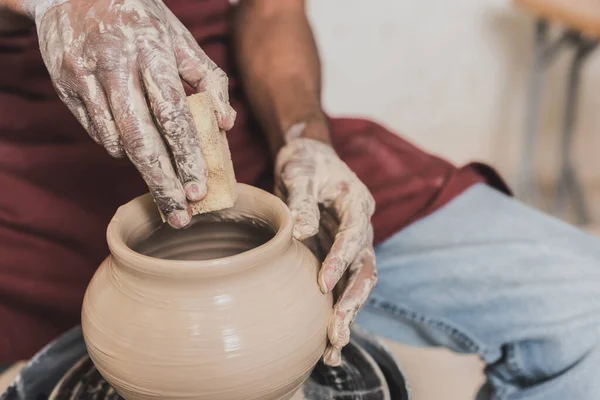 Close up view of young african american man holding sponge and making wet clay pot on wheel in pottery — Stock Photo