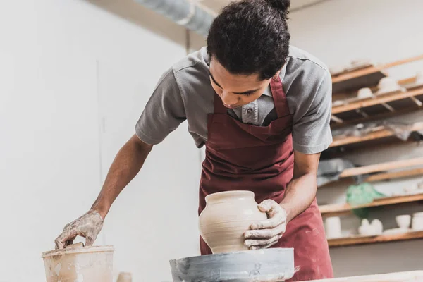 Молодий афроамериканський чоловік робить мокрий глиняний горщик на колесі і миє руку губкою в пластиковій коробці в кераміці — стокове фото