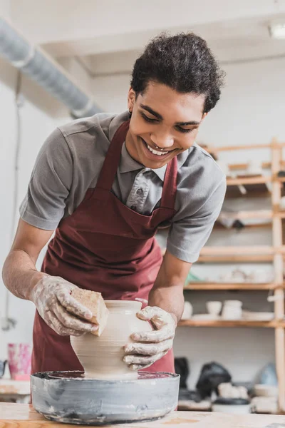 Sorridente giovane uomo africano americano che fa pentola di argilla bagnata su ruota con spugna in ceramica — Foto stock